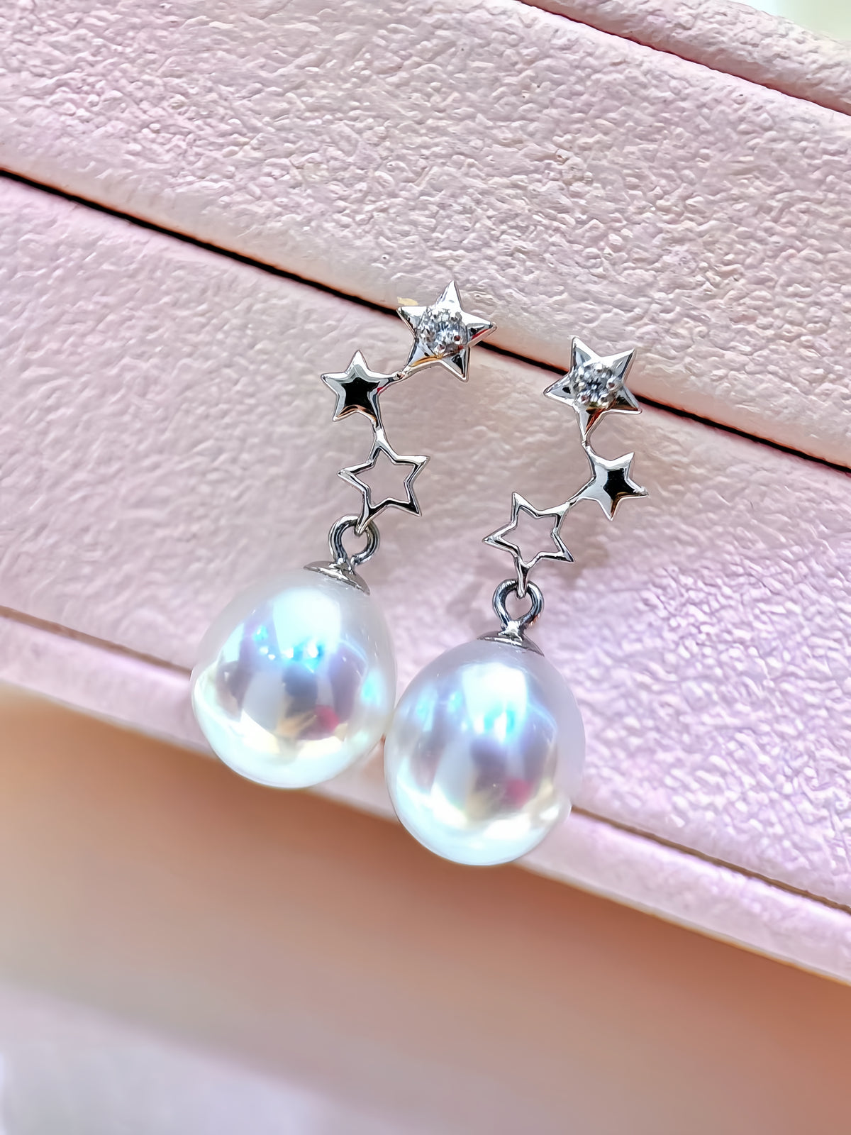 A pair of silver star drop earrings with large iridescent white pearls, displayed on a textured light pink jewelry box.
