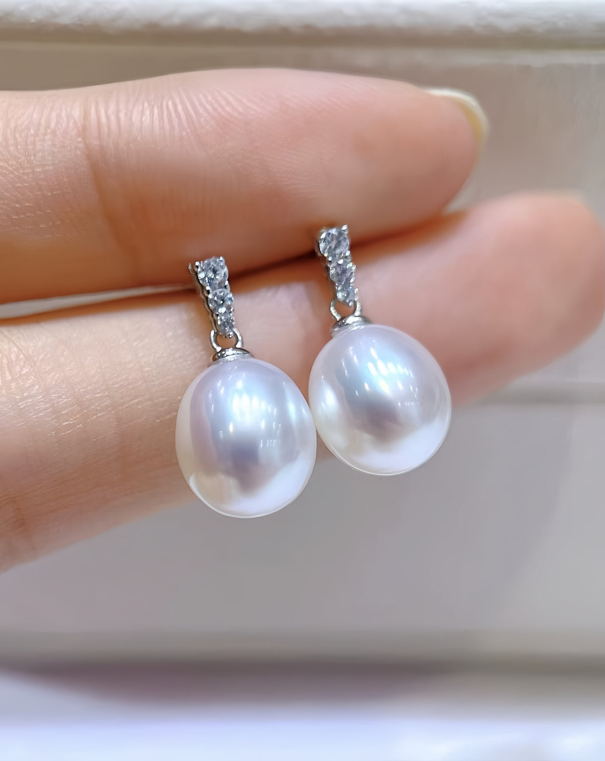 A pair of silver drop earrings with large white freshwater pearls and small clear stones at the top, held between fingers against a soft beige background.