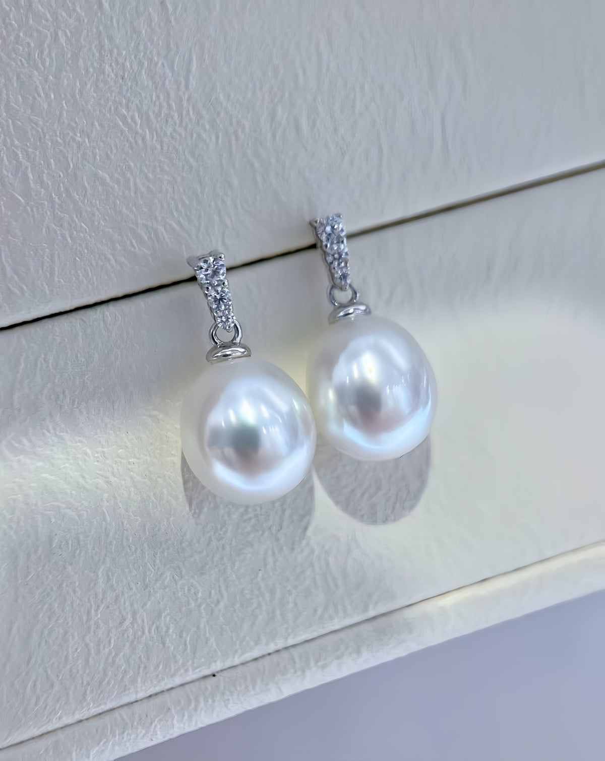 A pair of silver drop earrings with large, lustrous white freshwater pearls and small clear stones at the top, displayed on a textured cream-colored jewelry box.