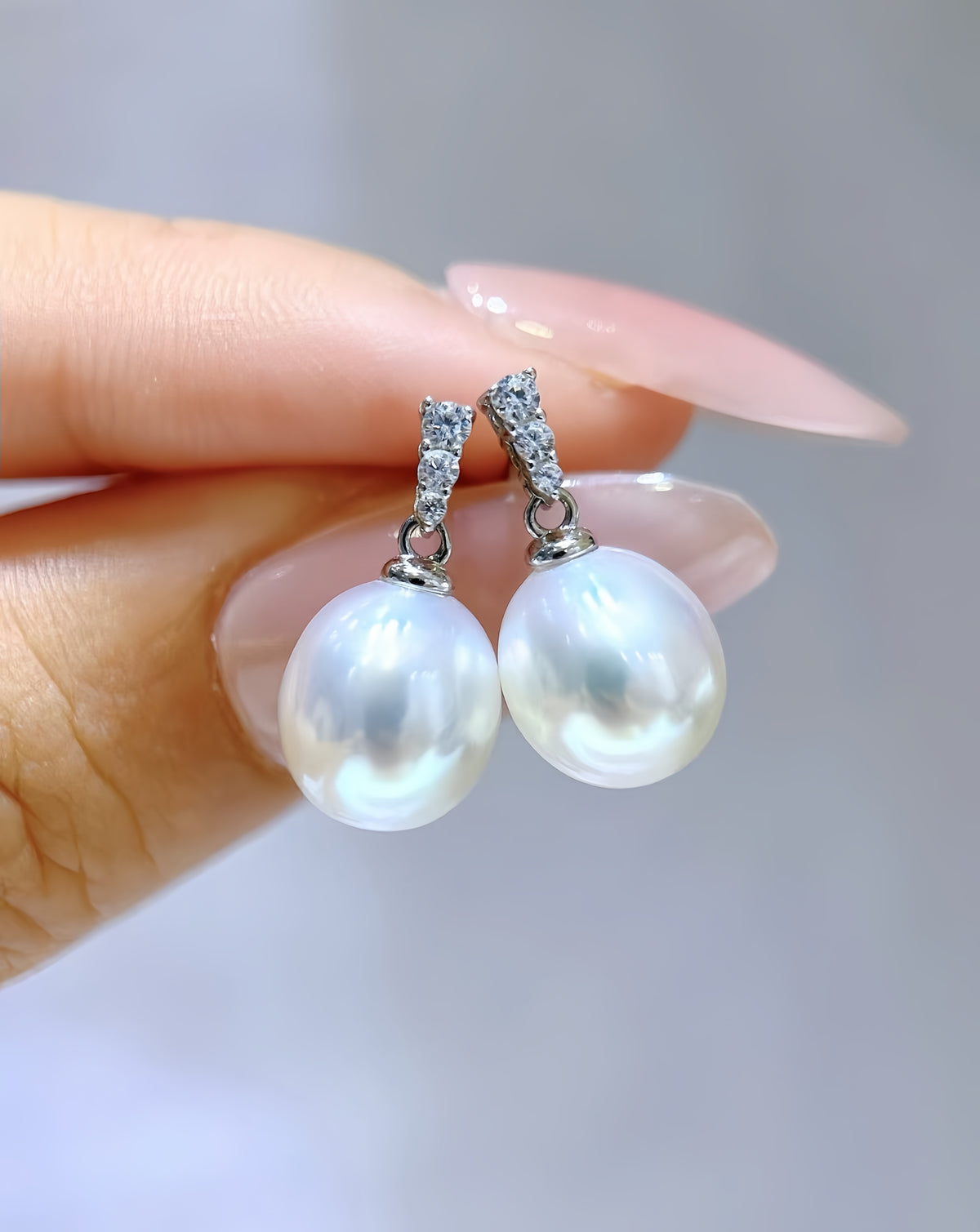 A close-up of a hand with long pinkish nails holding a pair of silver huggie earrings with large white freshwater pearls and small clear stones at the top.
