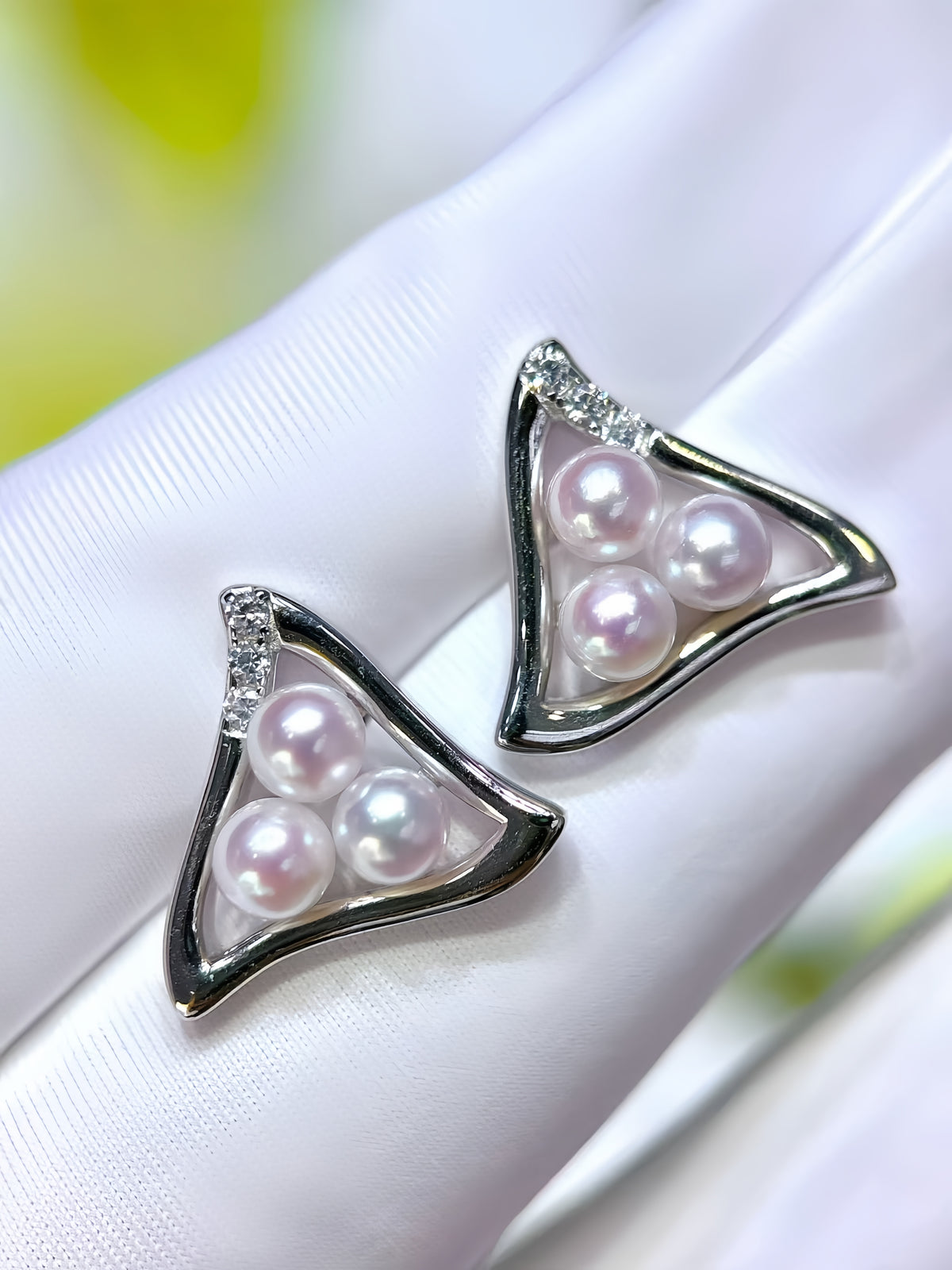 A pair of triangular stud earrings with three iridescent white pearls in each triangle, set in a polished silver-tone frame with a small clear crystal accent at the top, resting on a white textured surface.