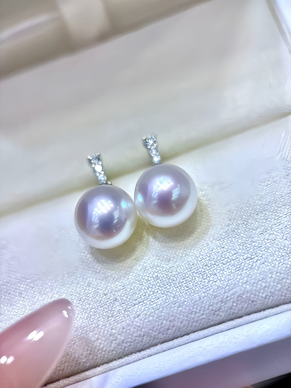 A pair of freshwater pearl stud earrings with silver-toned bezel settings, resting on a cream-colored jewelry display box. A pink manicured fingernail is visible in the bottom-left corner.