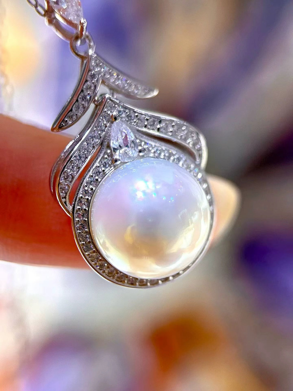 A close-up of a silver pendant necklace with a large iridescent white pearl and small clear stones, held between fingers against a blurred colorful background.